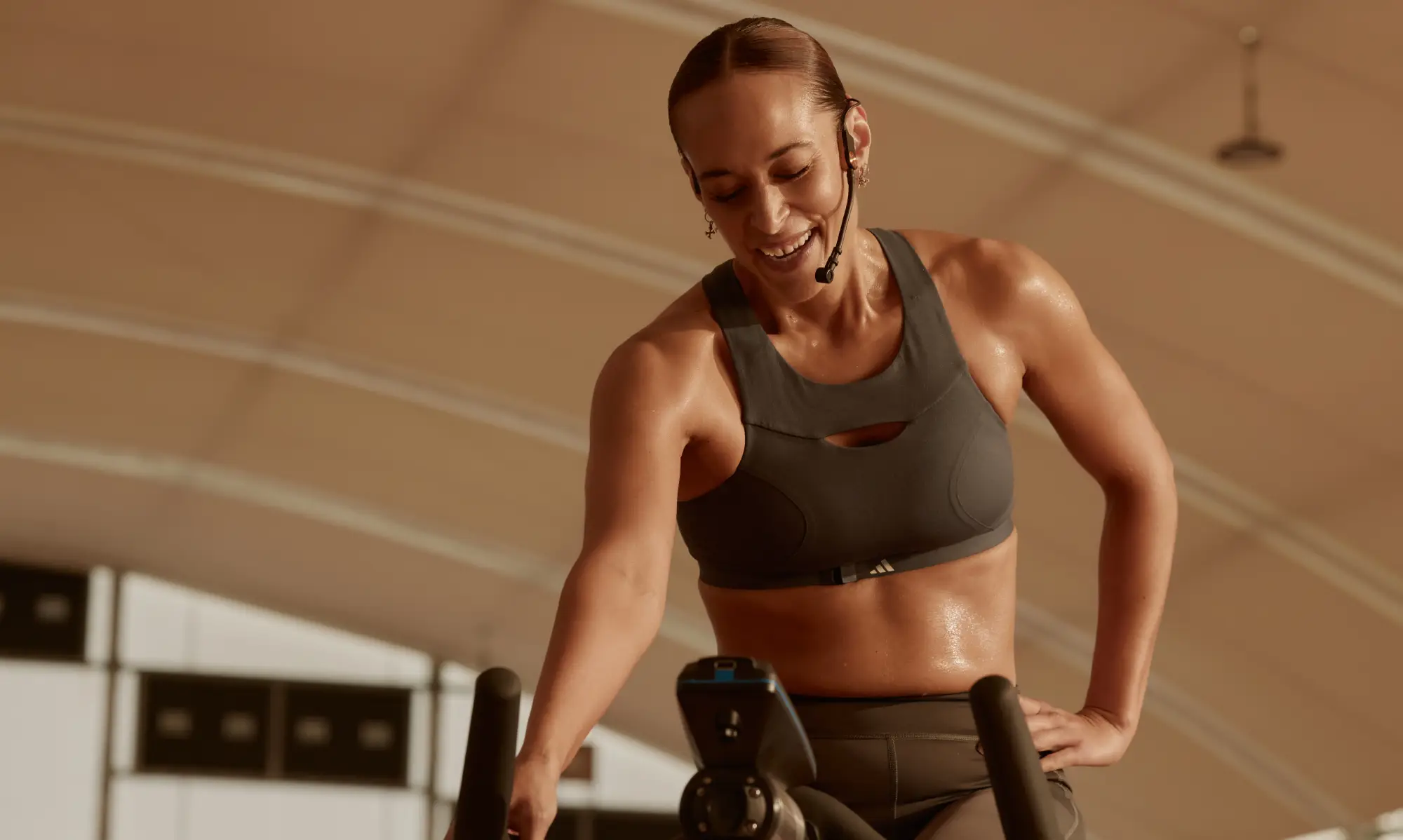 One person in a black sports bra and leggings exercises on a stationary bike in a gym with beige walls and ceiling. Gym equipment is visible in the background.